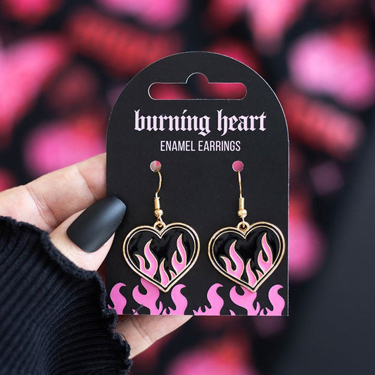 A hand with black matte nails holds a card showing pink and black flame heart earrings—ideal for Y2K style and rebellious outfits. The card reads burning heart ENAMEL EARRINGS and has pink flames along the bottom.