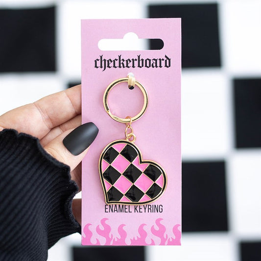 A hand with matte black nails holds a pink card labelled “checkerboard”, displaying a Y2K checkerboard heart keyring edged in gold. The background stands out with a bold black and white chequered pattern.