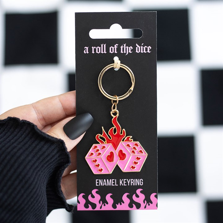 A hand with matte black nails holds a flaming dice keyring—two pink dice featuring red hearts and flames—on black packaging that reads A Roll of the Dice and ENAMEL KEYRING, set against a chequered background.