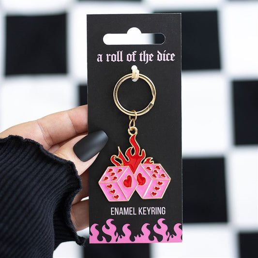 A hand with matte black nails holds a flaming dice keyring—two pink dice featuring red hearts and flames—on black packaging that reads A Roll of the Dice and ENAMEL KEYRING, set against a chequered background.