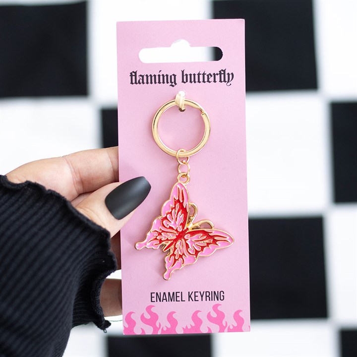 A hand with black matte nails holds a pink card featuring a gold and pink enamel cybersigil butterfly keyring. The card says flaming butterfly and ENAMEL KEYRING, pop-punk flame artwork at the bottom. Black-and-white background.