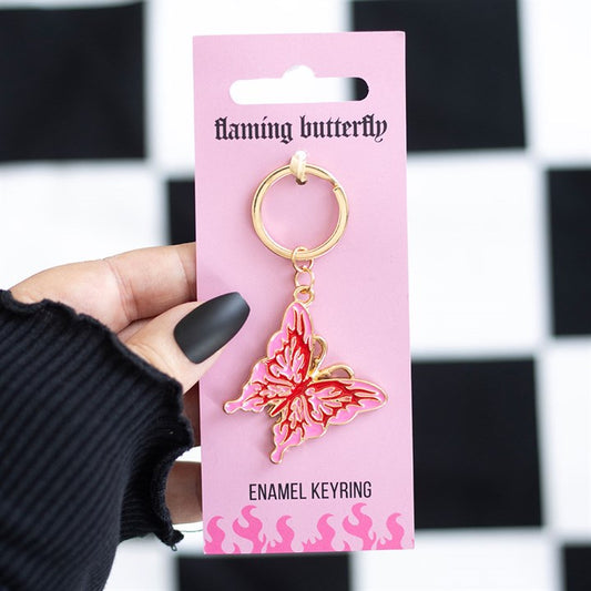 A hand with black matte nails holds a pink card featuring a gold and pink enamel cybersigil butterfly keyring. The card says flaming butterfly and ENAMEL KEYRING, pop-punk flame artwork at the bottom. Black-and-white background.