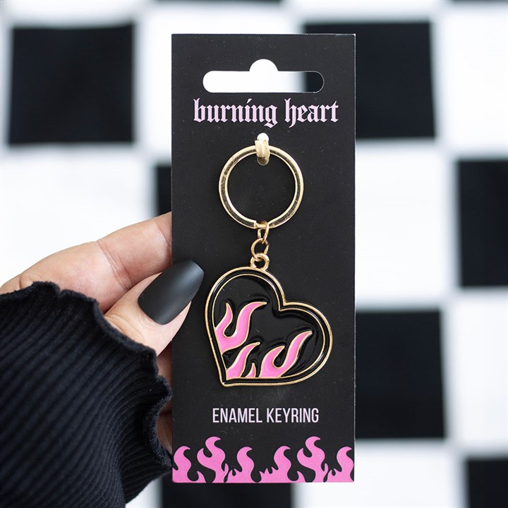 A hand with matte black nails holds a flaming heart keyring, its pink and black flames giving off rebellious energy, attached to bold packaging against a chequerboard background—ideal as a statement rucksack accessory.