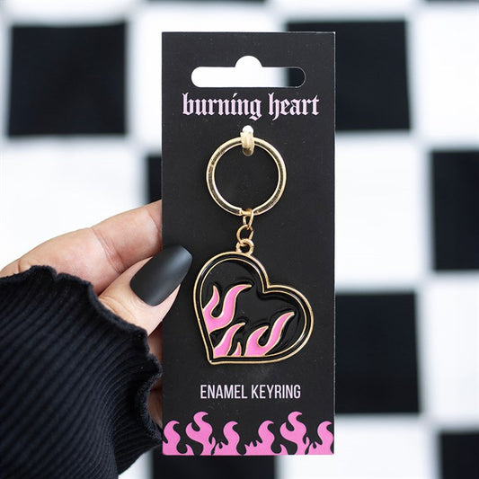 A hand with matte black nails holds a flaming heart keyring, its pink and black flames giving off rebellious energy, attached to bold packaging against a chequerboard background—ideal as a statement rucksack accessory.