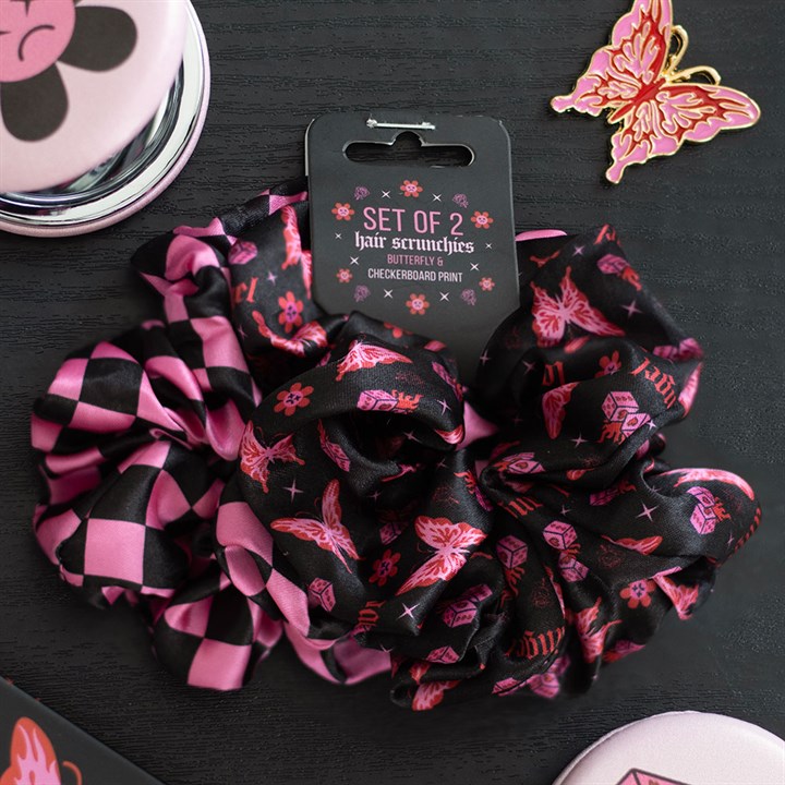 A set of two alternative scrunchies—one featuring a cybersigil butterfly print with flowers, the other in black and pink chequerboard—rests on a dark surface beside a butterfly pin, compact mirror, and pink-lidded box.