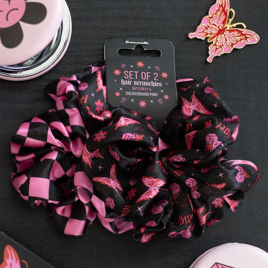 A set of two alternative scrunchies—one featuring a cybersigil butterfly print with flowers, the other in black and pink chequerboard—rests on a dark surface beside a butterfly pin, compact mirror, and pink-lidded box.
