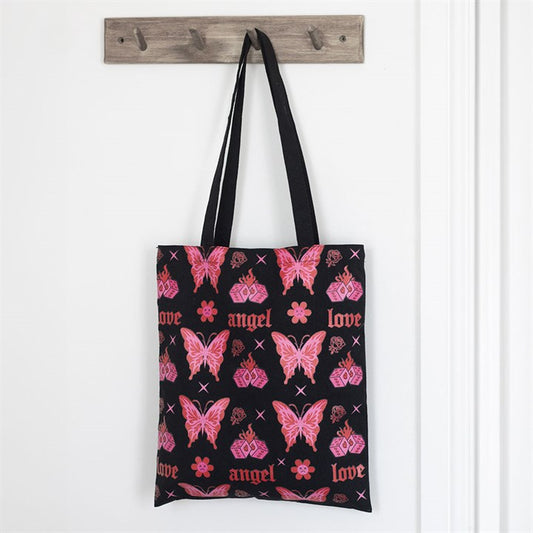A black, edgy tote bag with two handles hangs on a wooden hook, adorned with a pink Y2K nostalgia pattern of butterflies, hearts, flowers, “love”, and “angel”. The background shows a white wall and door.