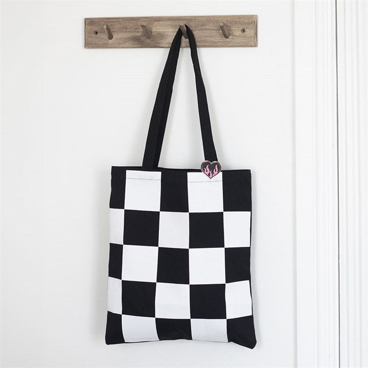 A chequerboard tote bag with two black handles hangs from a wooden wall rack, adorned with a small pink and black heart pin badge that adds pop-punk flair near the top right corner.