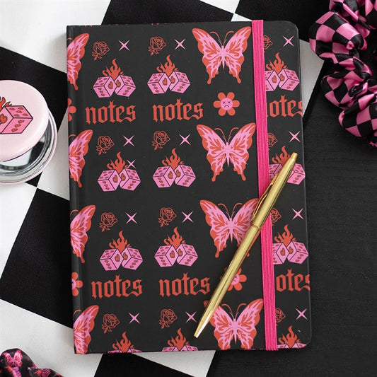 A Y2K-inspired A5 notebook with pink and red designs—dice, butterflies, flames, flowers, and “notes”—rests on a black-and-white chequered surface with a gold pen, scrunchie, and dice-shaped container nearby.