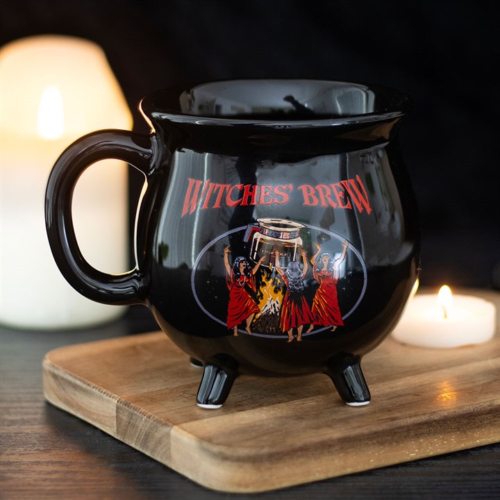 A Witches’ Brew cauldron mug, labelled in red, depicts three witches dancing around a fire. This witch’s coffee mug rests on a wooden board beside two lit candles, set against a dark background.