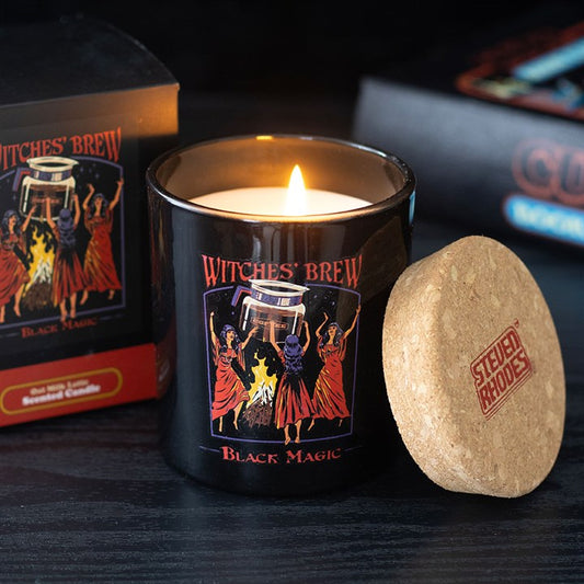 A black candle labelled ‘Witches’ Brew Black Magic’ featuring artwork of three witches dancing around a cauldron. This oat milk latte-scented candle by Steven Rhodes is lit beside its box and cork lid on a dark wooden surface.