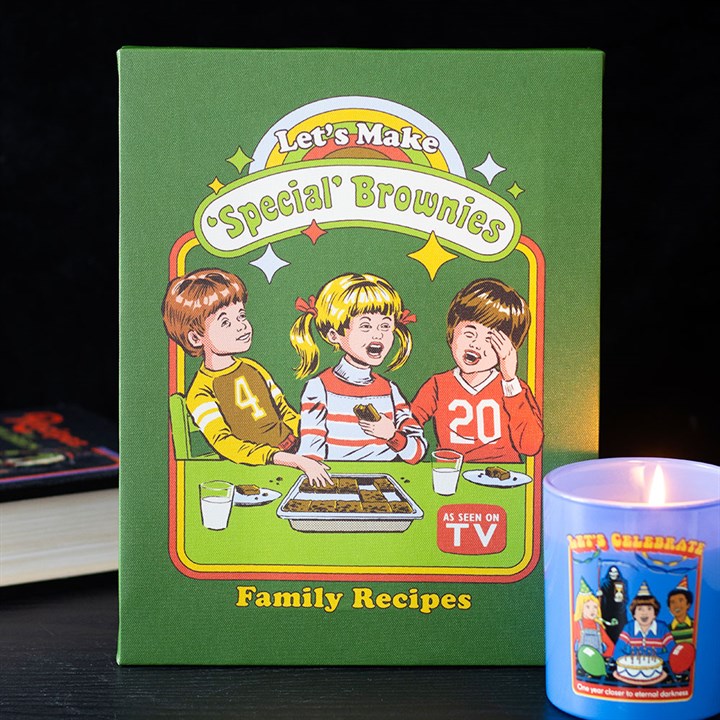 A green recipe book titled Let’s Make Special Brownies: Family Recipes features a cartoon of three children eating brownies. A stretched canvas plaque and retro gifts add charm, with a lit candle and a book in the background.