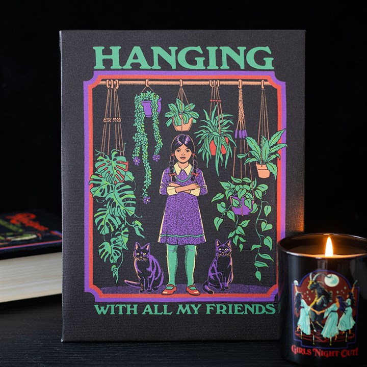 A candle burns next to a book and retro wall art depicting a girl with long dark hair among hanging and potted plants, flanked by two black cats. The text on the artwork reads: HANGING WITH ALL MY FRIENDS.