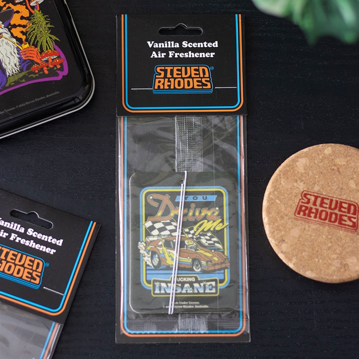 A packaged Steven Rhodes air freshener with a vanilla scent lies on a dark surface near a cork coaster and metal tin. The retro race car design and “Driving Me Insane” text make it perfect for fans of quirky, retro gifts.