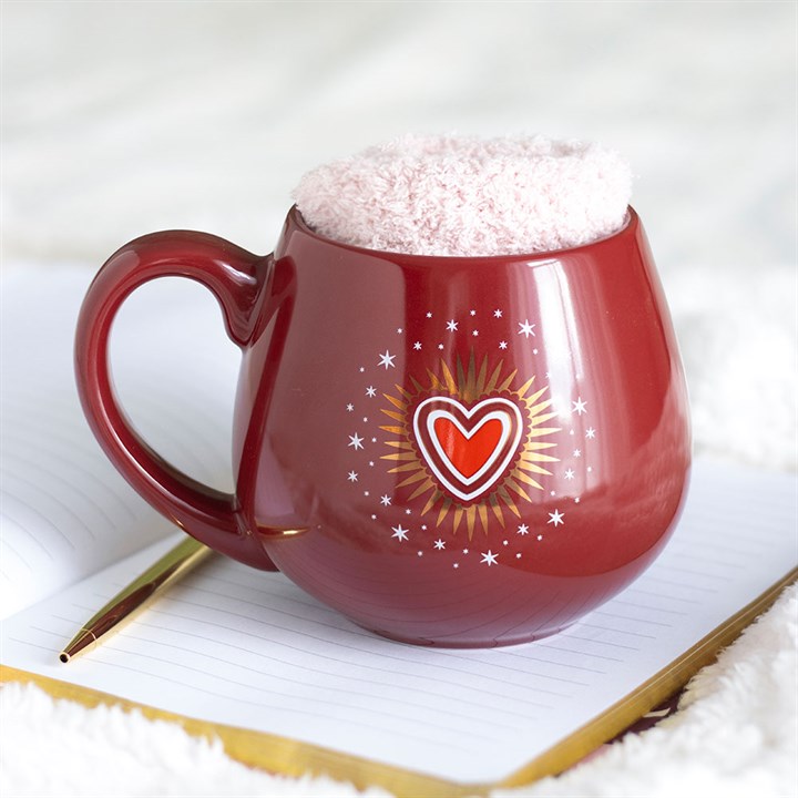 A red mug featuring a heart and starburst design rests on an open notebook alongside a gold pen. Inside the mug is an unidentified, soft pink item—perhaps socks or a cosy cloth—set against a soft white background.
