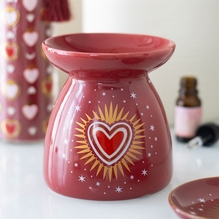 A red ceramic oil burner featuring a radiant heart motif, golden rays, and white stars. In the background, a bottle and another item adorned with delightful heart patterns—ideal for your favourite fragrance oil.