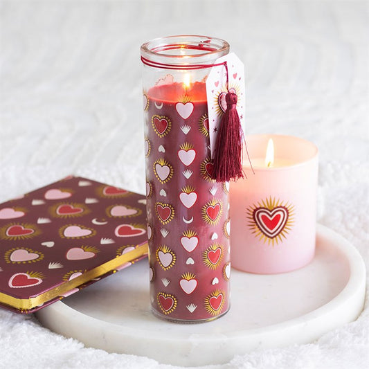 A tall red sacred heart candle with gold, white, and pink heart motifs and a tassel tag burns on a white tray—ideal Valentines Day decor—next to a pink radiant heart candle and a coordinating heart-patterned notebook.