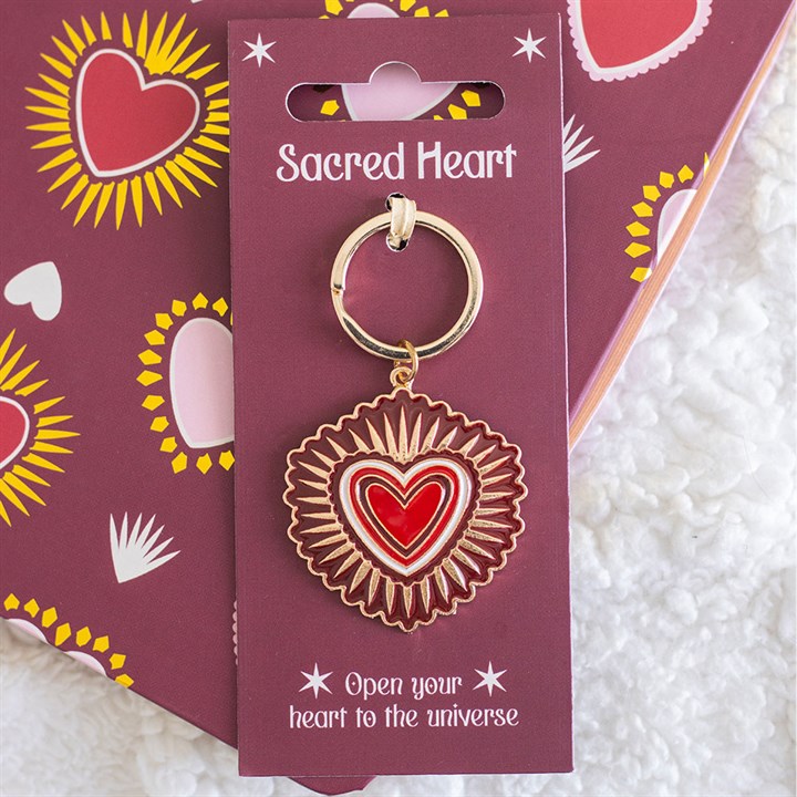 A gold-accented sacred heart keyring with a red and white design is attached to a purple card reading “Sacred Heart”—a thoughtful Valentine’s Day gift on a matching heart-patterned background.