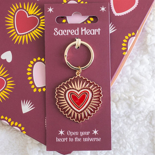 A gold-accented sacred heart keyring with a red and white design is attached to a purple card reading “Sacred Heart”—a thoughtful Valentine’s Day gift on a matching heart-patterned background.