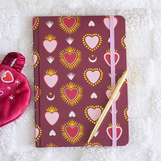 A maroon A5 notebook with colourful heart patterns, fastened by a pink elastic band, rests on a white textured blanket. A gold pen lies diagonally across the notebook, and a red heart-patterned pouch peeks in from the left.