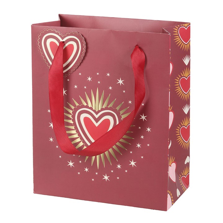 A love-themed gift bag with red handles, gold and white stars, and several heart motifs, including a prominent sacred heart encircled by golden rays. A heart-shaped gift tag dangles from one handle.