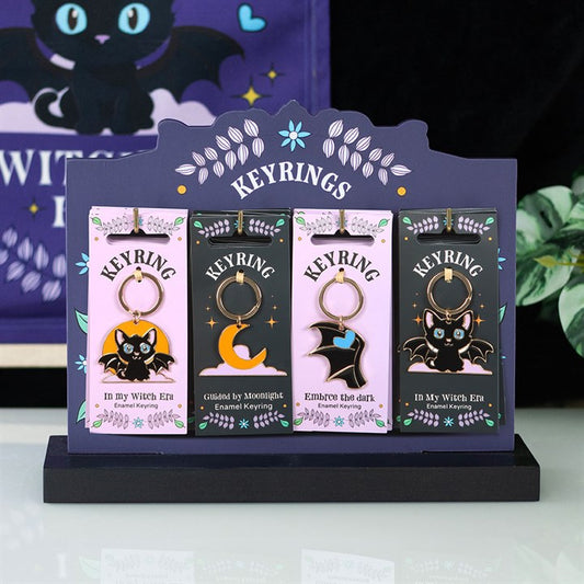 A display stand holds four witch-themed enamel keyrings, each featuring a bat or moon design—perfect for lovers of gothic aesthetics. The purple background banner is adorned with a black cat and stars.