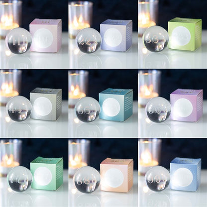 A 3x3 grid of spherical candles sits alongside matching square boxes in pastel hues, each pair adorned with laser-engraved numbers from 08 to 99. Blurred candles in glass holders create a warm, cosy ambience, reminiscent of angel numbers guiding through a gentle glow.