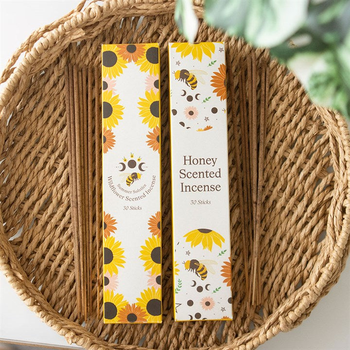 A woven basket holds two boxes of incense sticks—one with a wildflower scent and the other honey-scented—decorated with sunflowers, bees, and stars. Green leaves peek out above the vibrant packaging.