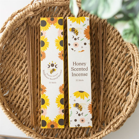 A woven basket holds two boxes of incense sticks—one with a wildflower scent and the other honey-scented—decorated with sunflowers, bees, and stars. Green leaves peek out above the vibrant packaging.