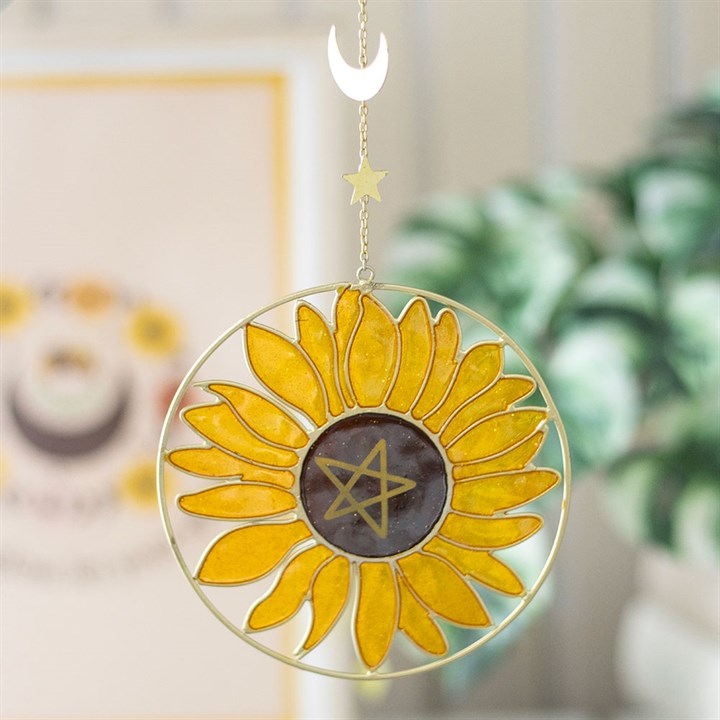 A sunflower suncatcher with a black pentagram centre radiates a cheerful glow. Hanging from a gold ring, crescent moon, and star, it captures the spirit of the Summer Solstice against a blurred leafy backdrop.
