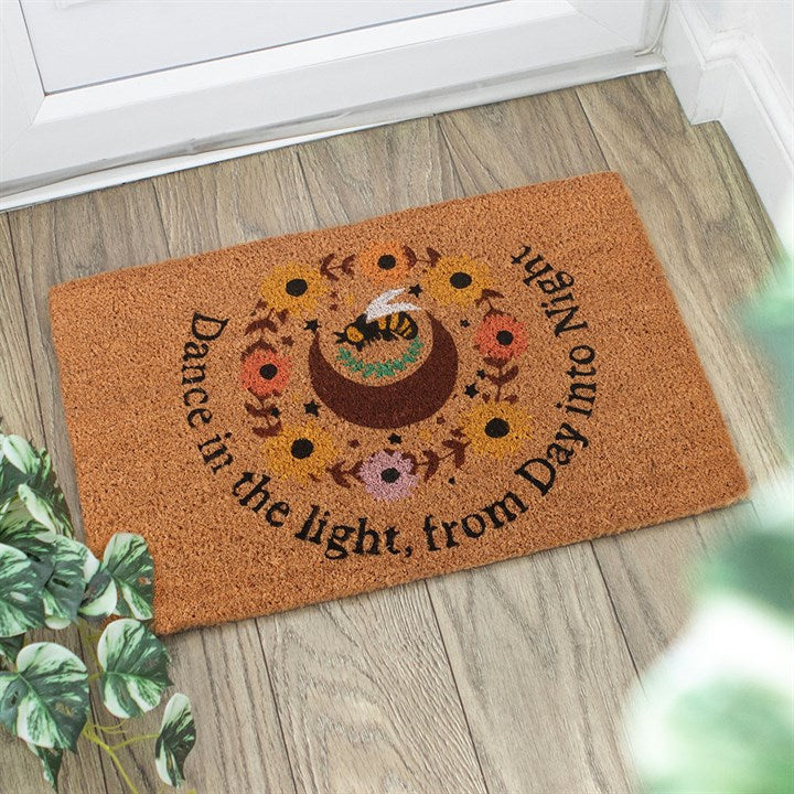 A Summer Solstice doormat featuring a bee on a crescent moon, surrounded by flowers and foliage. “Dance in the light, from Day into Night.” The perfect rustic entrance hall décor beside leafy plants on a wooden floor.