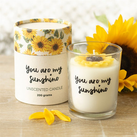 A decorative unscented candle in a glass holder labelled “You are my sunshine” sits beside a sunflower-themed box. A sunflower petal adorns the top, with a sunflower and loose petals in the background.