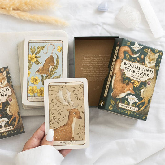 A hand holds a tarot card featuring an illustration of a deer and oak leaves—part of the Woodland Wardens oracle deck. Nearby, more animal-and-plant pairings and crystals are arranged beside an open box by Jessica Roux on a white surface.