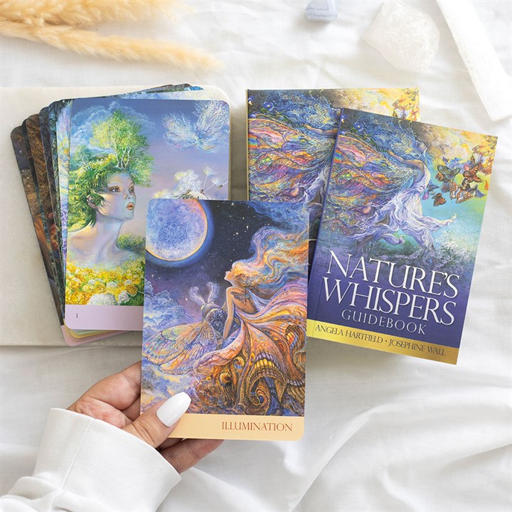 A hand holds an “Illumination” card from vibrant nature oracle cards. Other ethereal designs, inspired by Josephine Wall’s artwork, and the “Nature’s Whispers Guidebook” rest on a white cloth background.
