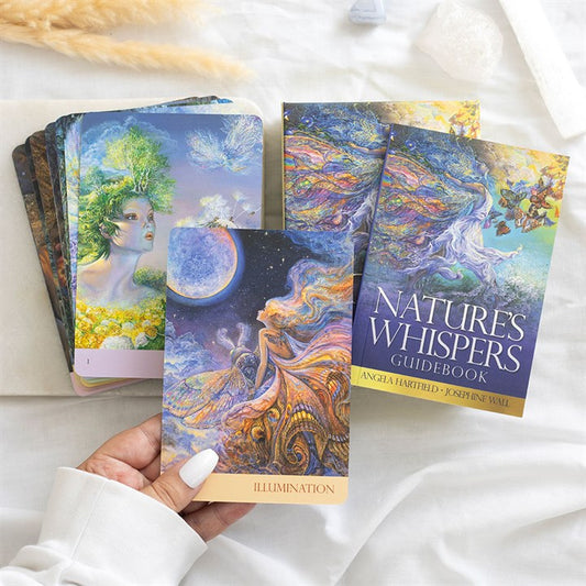 A hand holds an “Illumination” card from vibrant nature oracle cards. Other ethereal designs, inspired by Josephine Wall’s artwork, and the “Nature’s Whispers Guidebook” rest on a white cloth background.