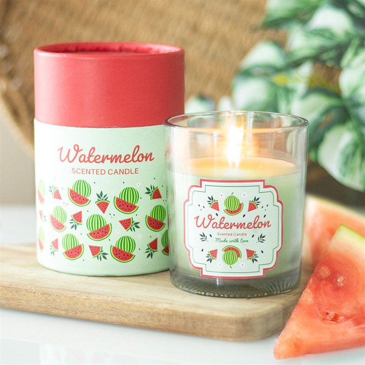A lit watermelon-scented candle with a wooden lid sits beside its decorative box, both featuring a fruity watermelon print; slices of watermelon and leafy greens are visible in the background.