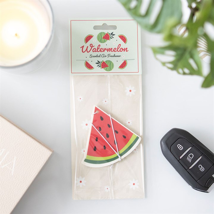 A watermelon air freshener shaped like a juicy slice sits beside a car key, lit candle, pale pink book and green leaves, filling the area with a fruity aroma. The label reads “Watermelon Scented Air Freshener.”.