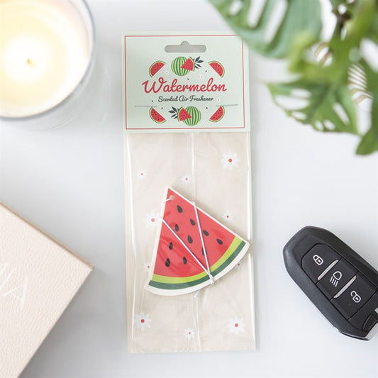 A watermelon air freshener shaped like a juicy slice sits beside a car key, lit candle, pale pink book and green leaves, filling the area with a fruity aroma. The label reads “Watermelon Scented Air Freshener.”.