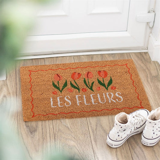 A brown tulip doormat with red floral patterns and LES FLEURS in white rests on a wooden floor by a white door; ideal for garden lovers, with starry trainers beside it and greenery blurred in the foreground.