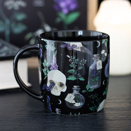A gothic mug adorned with white skulls, potion bottles, purple flowers, green plants, and crescent moons rests on a dark tabletop—an artistic design by Venom & Vines. A blurry book and candle glow in the background.