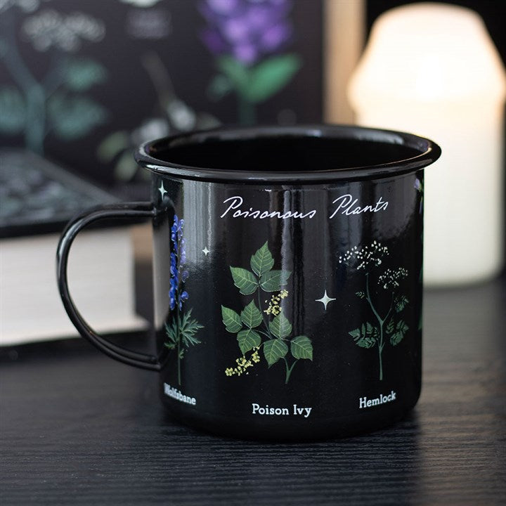 A gothic mug labelled Poisonous Plants, featuring illustrations of nightshade, poison ivy, and hemlock, sits on a dark surface with a book and a softly glowing candle in the blurred background.