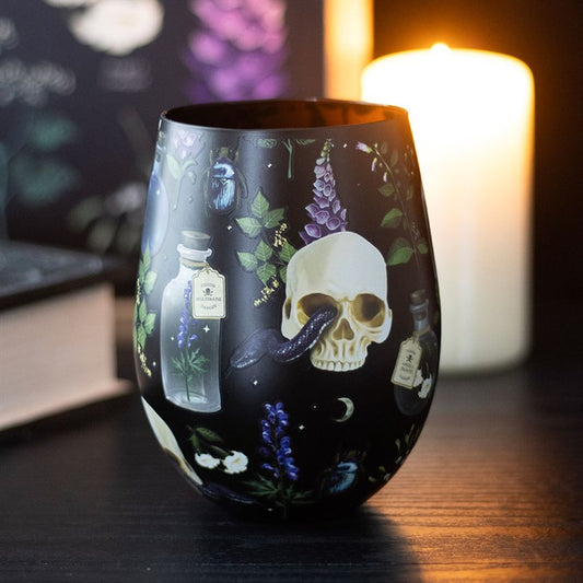 A black stemless glass, adorned with a gothic skull, purple and blue flowers, potion bottles, and a black snake, sits on a dark table. A lit candle and a closed book in the background contribute to the moody atmosphere.