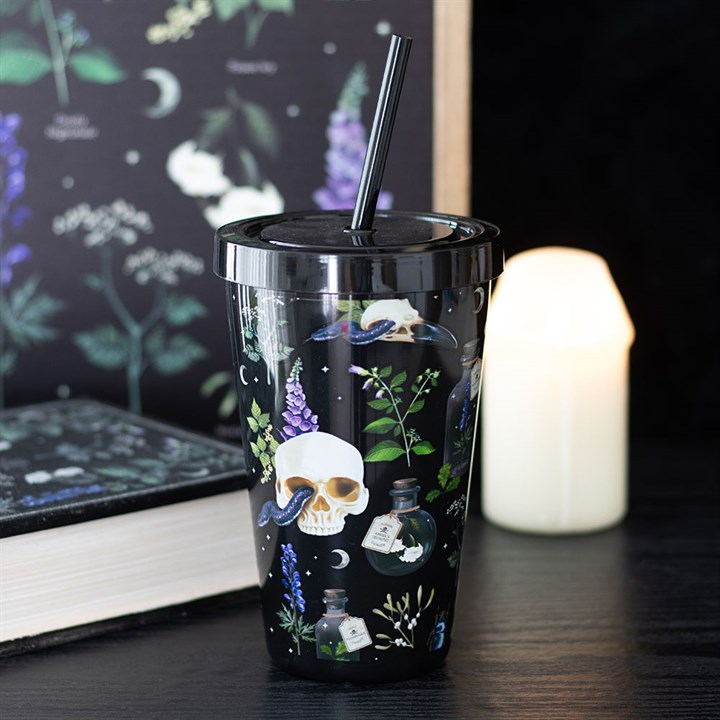 A reusable plastic tumbler featuring a design of a skull and poisonous plants, complete with lid and straw, sits stylishly on a dark table among a white candle, a book, and a botanical-themed backdrop—functional yet fashionable accessories.