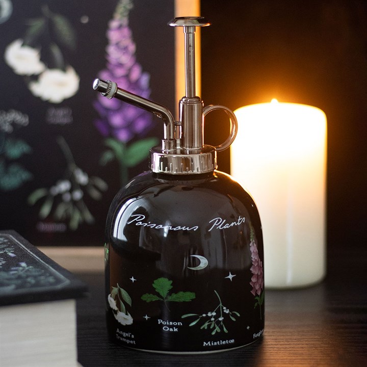 A black glass plant mister labelled ‘Poisonous Plants’ features botanical illustrations of toxic flora. It rests on a dark surface beside an open book and a lit white candle, making it perfect for any indoor garden.