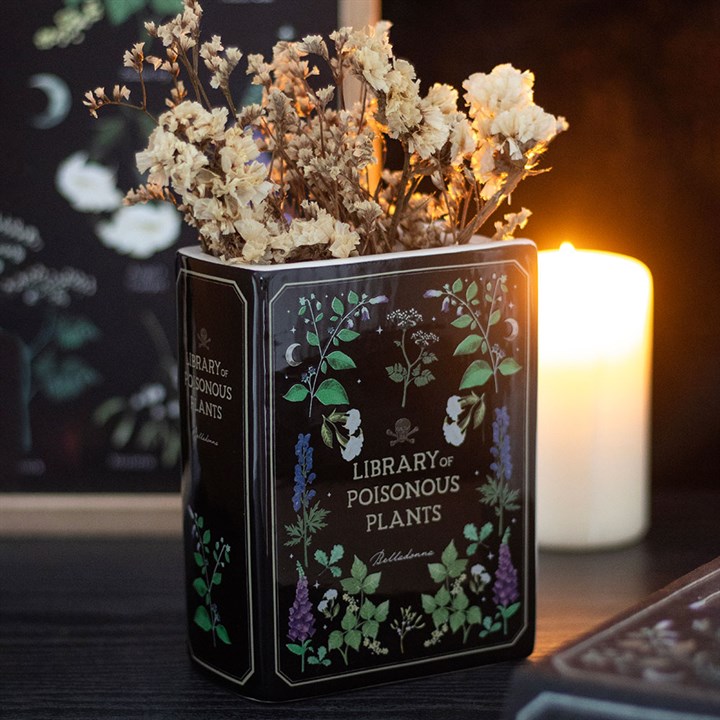 A decorative piece with dark elegance, this black, book-shaped vase labelled Library of Poisonous Plants holds dried flowers while a warm candle glows in the background on a dark surface.