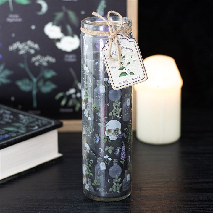 A tall glass tube candle with a deadly nightshade design—skulls, potion bottles, and herbs—features a twine bow and Nightshade label. Perfect gothic-inspired décor beside a glowing candle and book on a dark table.