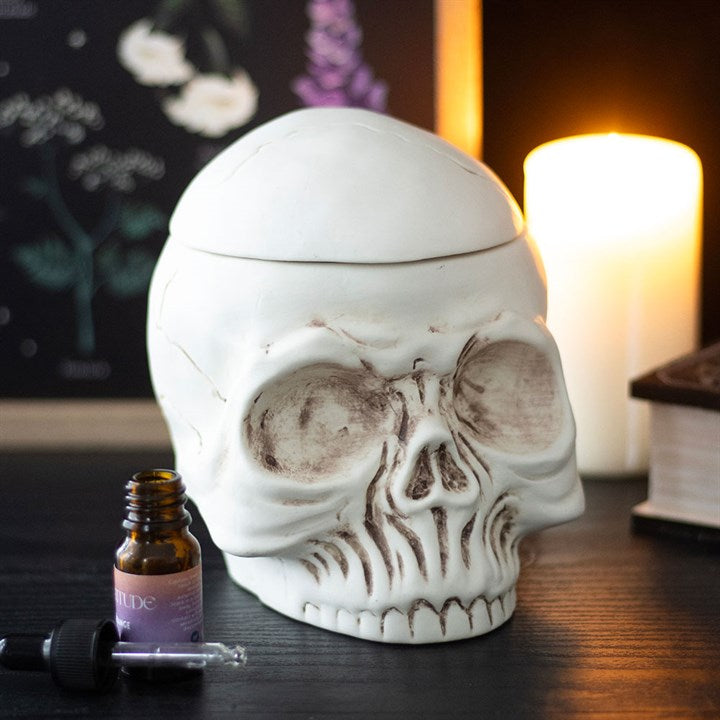 A white ceramic skull oil burner sits on a dark table, its lid slightly ajar. Nearby are a lit candle, a brown glass dropper bottle, a book, and a botanical print—perfect for enthusiasts of gothic décor.
