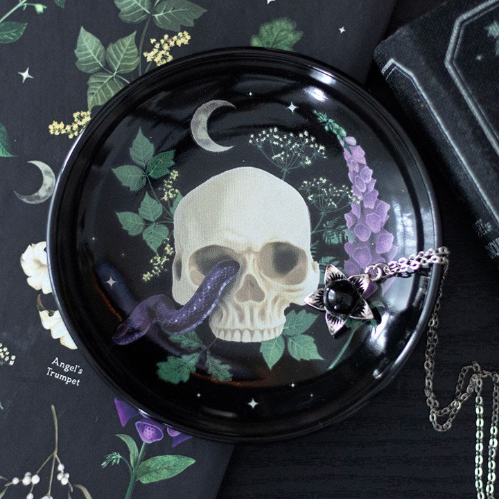 A ceramic dish in Gothic décor features a skull and snake motif, purple flowers, greenery, a crescent moon, and a silver pendant. Botanical illustrations and a book surround this striking piece.