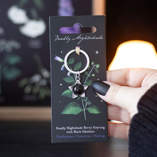 A hand with black nail varnish holds a gothic keyring—the “Deadly Nightshade Berry Keyring” from the Venom and Vines collection—adorned with a black obsidian charm, packaged on an illustrated nightshade card.