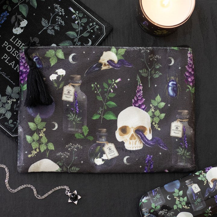 A gothic make-up bag decorated with skulls, potion bottles, purple flowers, moons and crows rests on a black surface beside beauty essentials—a candle, chain necklace and matching botanical print cloth.